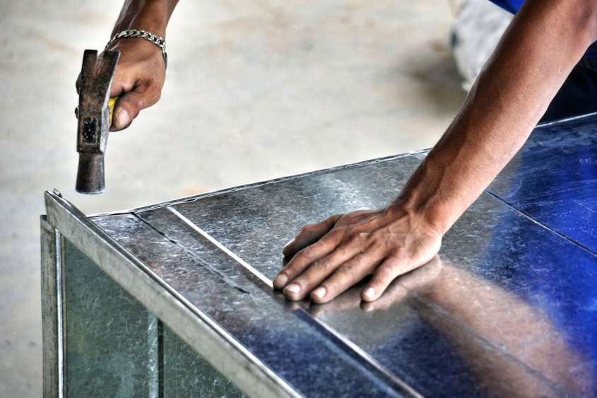 Eine Hand, die einem Hammer hält und damit das Blech eines Luftkanals bearbeitet
