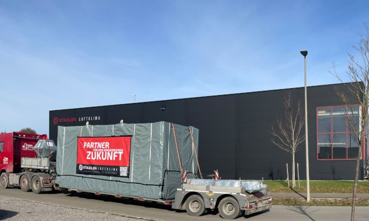Ein LKW transportiert ein verpacktes Lüftungsmodul ab. Auf der Verpackung ist ein Banner mit der Aufschrift "Partner für eine Emissionsfreie Zukunft" zu sehen, das die Stadler Luftklima bewirbt.