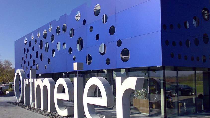 Bürogebäude mit Aluminiumfassade eloxiert Eine blaue Gebäudefassade, aus der kreisrunde Löcher geschnitten wurden. Davor in großen, weißen Lettern: Ortmeier