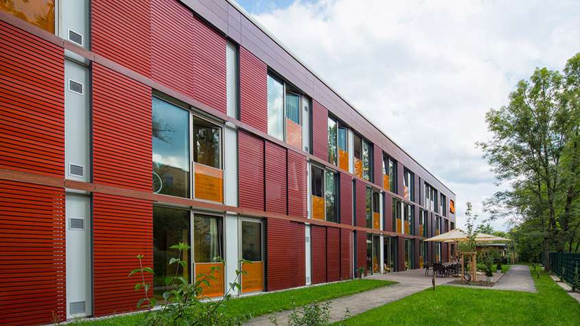 Seniorenheim mit Faserverbundplattenfassade Kempten Eine rote Fassade hinter einer grünen Wiese und einem Weg