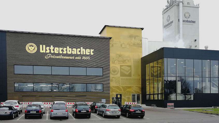 Aluminiumfassade eloxiert Ustersbach Gold-braune Gebäude hinter einem Parkplatz. Eine Fassade an der rechten Seite trägt die Aufschrift „Ustersbacher“