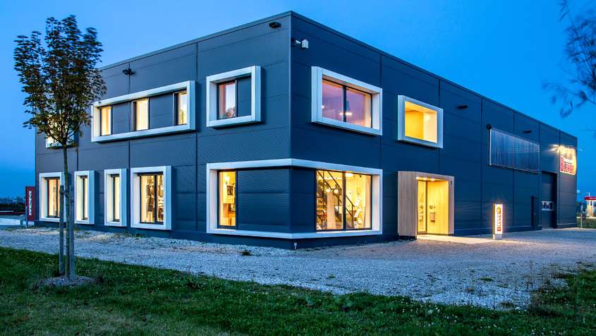 Büro und Lager mit Isopaneelen und Faserverbundplatten Ein rechteckiges Gebäude aus dunkelgrauen Paneelen mit beleuchteten Fenstern und breiten, weißen Fensterrahmen