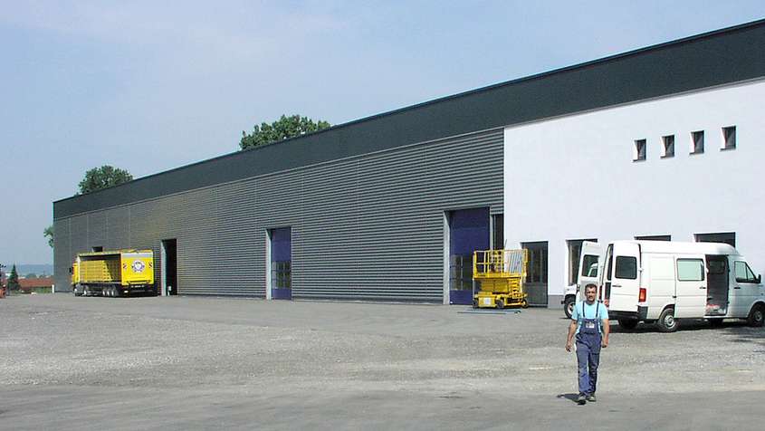 Fassade mit Aluminiumwelle und Faserverbundplatten Ustersbach Langes Gebäude mit Aluminiumfassade, einem gehenden Mann im Vorder- und einem gelben LKW im Hintergrund