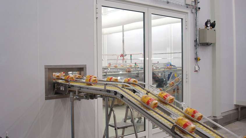 GLASBORDTRENNWAND AUF HYGIENESOCKEL WEISSENFELS Ein Fließband, das abgepacktes Brot durch eine weiße Wand transportiert, die durch ein großes Fenster durchbrochen wird, durch das man einen Raum mit mehr Fließbändern sehen kann