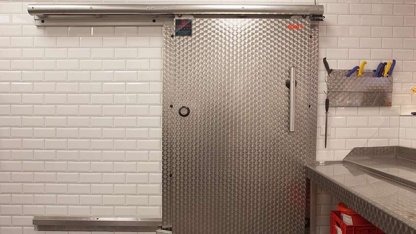 KÜHLRAUMSCHIEBETÜR IN DER FLEISCHVORBEREITUNG / SIMMEL-MARKT Einzelne Schiebetür aus Metall vor einer weißen Backsteinwand