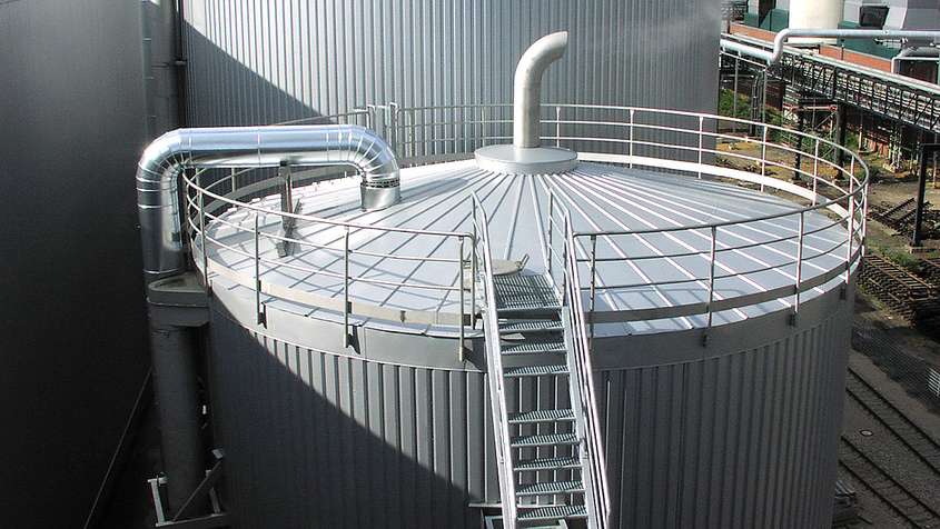 THERMISCHE ISOLIERUNG WASSERTANK HÜRTH Ein großes Silo mit einer Treppe aus Metall. Etwas Dampf ströhmt aus einem Rohr an der Spitze des Silos