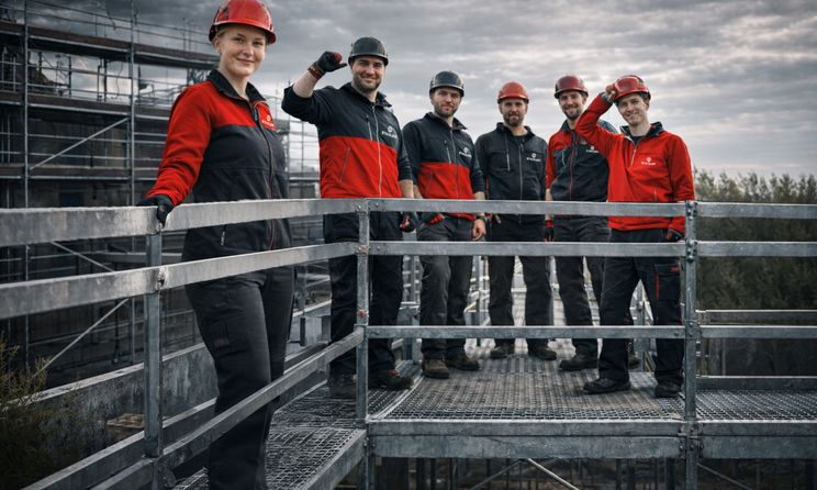 Eine Frau und fünf Männer in Arbeitskleidung an einem Baugerüst